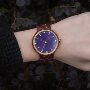 An elegant women's wooden watch featuring a purple dial, gold-tone accents, diamond-like details, and a two-tone wood and metal band, showcased on a wrist against a backdrop of dark foliage to highlight its luxurious and natural design.