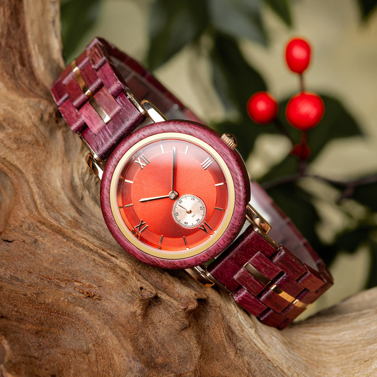 A stylish women's wooden watch featuring a red dial, Roman numerals, sub-dial, and gold-accented details, presented against a backdrop of rustic wood and green foliage to highlight its sophisticated and vibrant aesthetic.
