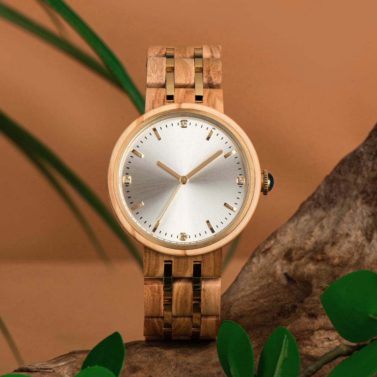 An elegant women's wooden watch featuring a silver dial, gold-tone accents, diamond-like details, and a two-tone wood and metal band, presented on rustic wood with green foliage to highlight its luxurious and natural design.