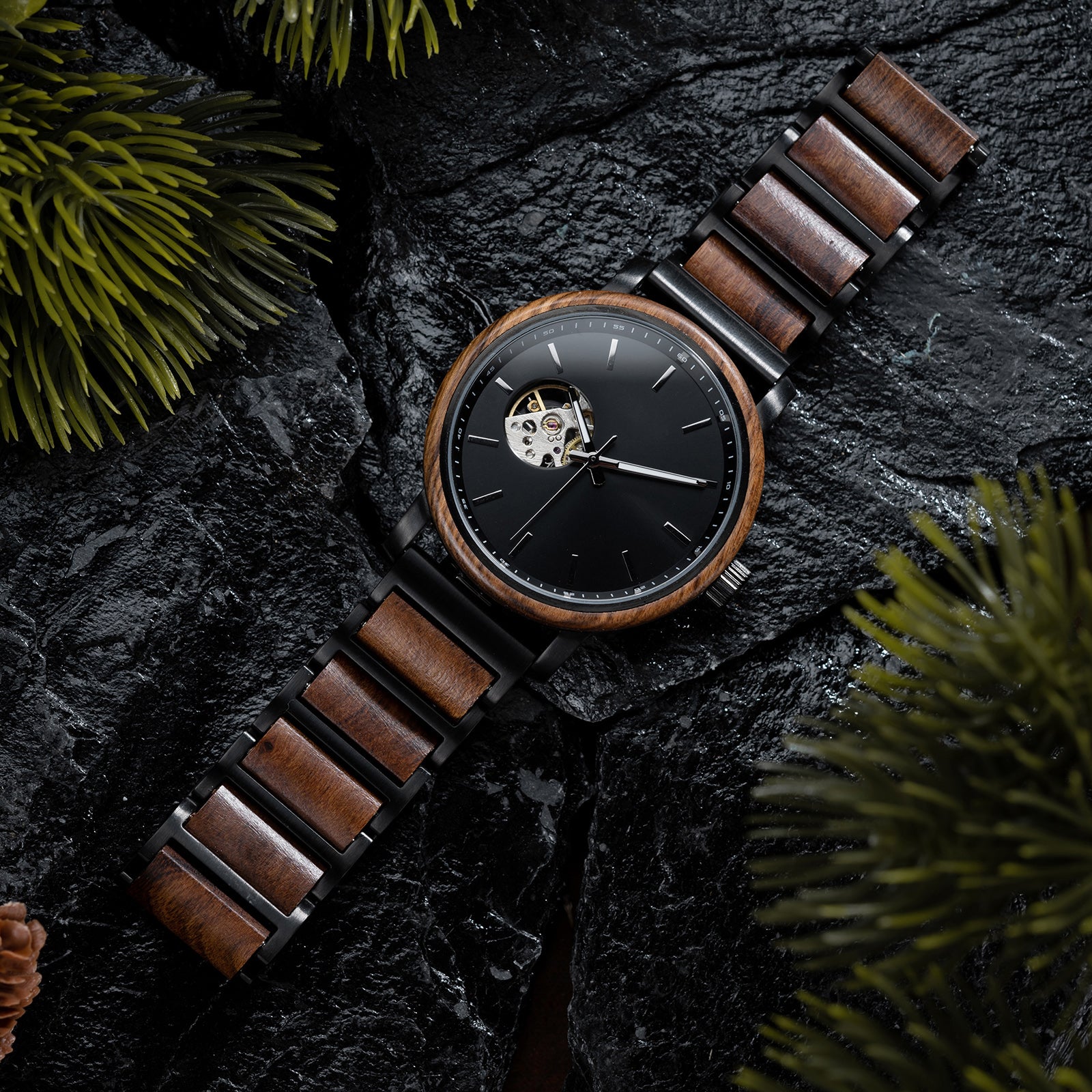 A stylish skeleton automatic watch with a wooden bezel, black metal and wood combined band, featuring an exposed movement on a black dial, placed on dark rocks surrounded by green foliage.