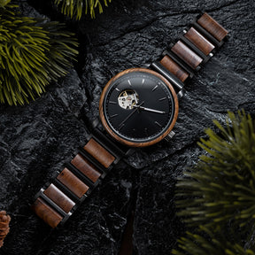 A stylish skeleton automatic watch with a wooden bezel, black metal and wood combined band, featuring an exposed movement on a black dial, placed on dark rocks surrounded by green foliage.