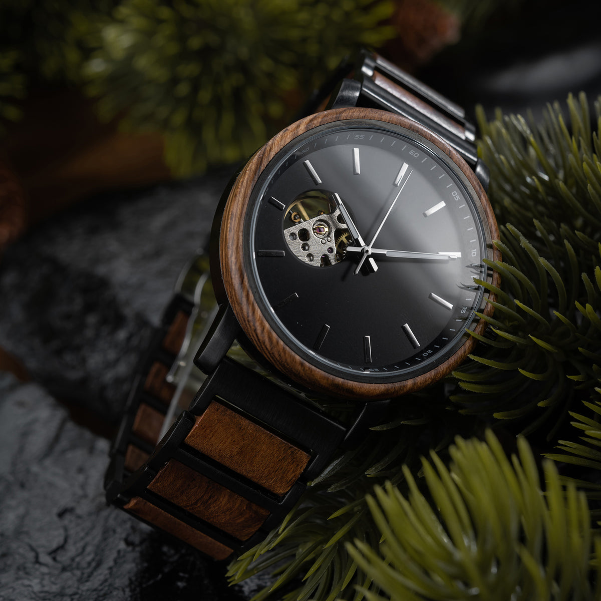 A stylish automatic watch with a wooden bezel, black metal and wood combined band, featuring a skeleton movement window on a black dial, placed among green foliage and dark rocks.