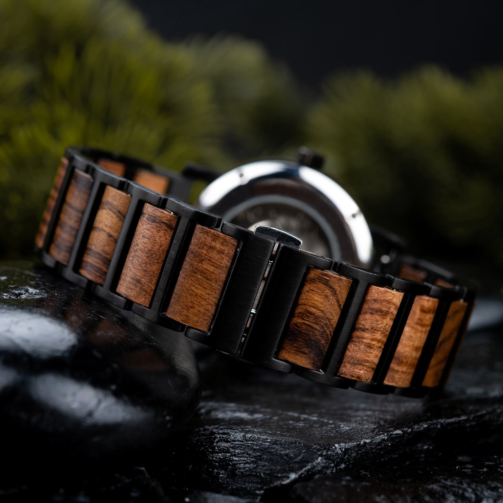 A close-up view of a watch band combining black metal and wooden links, placed on dark rocks with a blurred green foliage background.