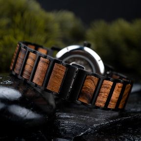 A close-up view of a watch band combining black metal and wooden links, placed on dark rocks with a blurred green foliage background.