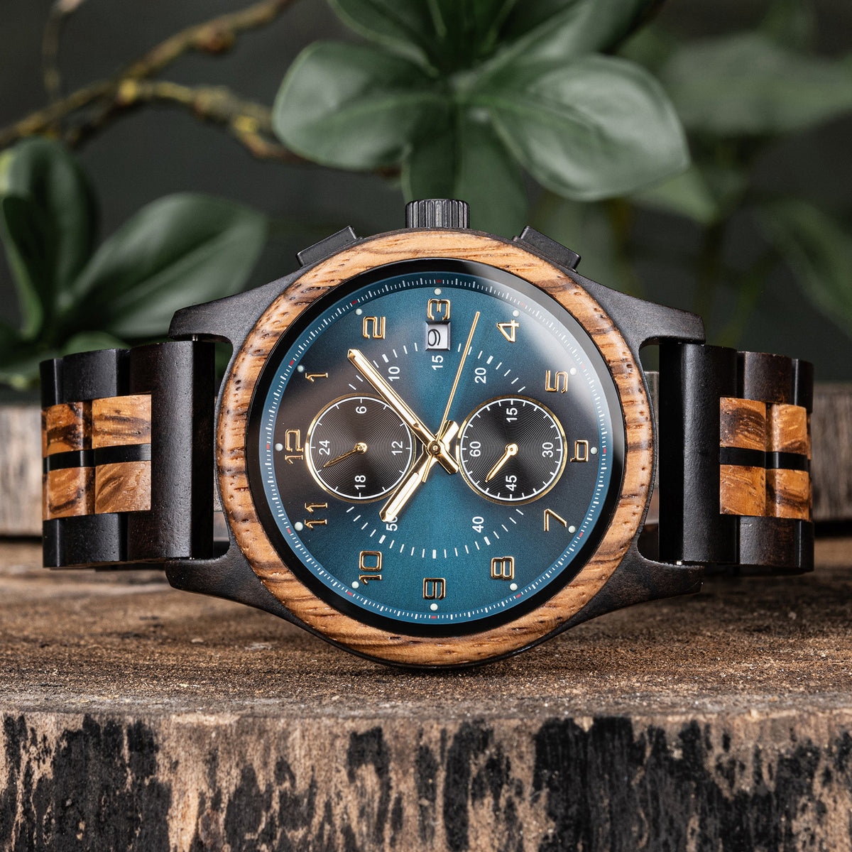 A men's chronograph watch with wooden and black metal accents, featuring a teal dial with gold-tone hands and markers, displayed on a rustic wooden surface with green foliage in the background.
