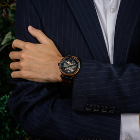 A men's skeleton mechanical watch with a wooden and black metal case and band, worn on the wrist of a person dressed in a navy pinstripe suit and white dress shirt.