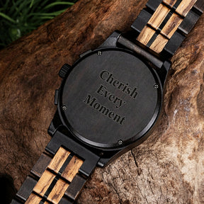 The back of a men's watch with a combination of dark wood and black metal band, placed on a textured piece of wood with green foliage in the background.