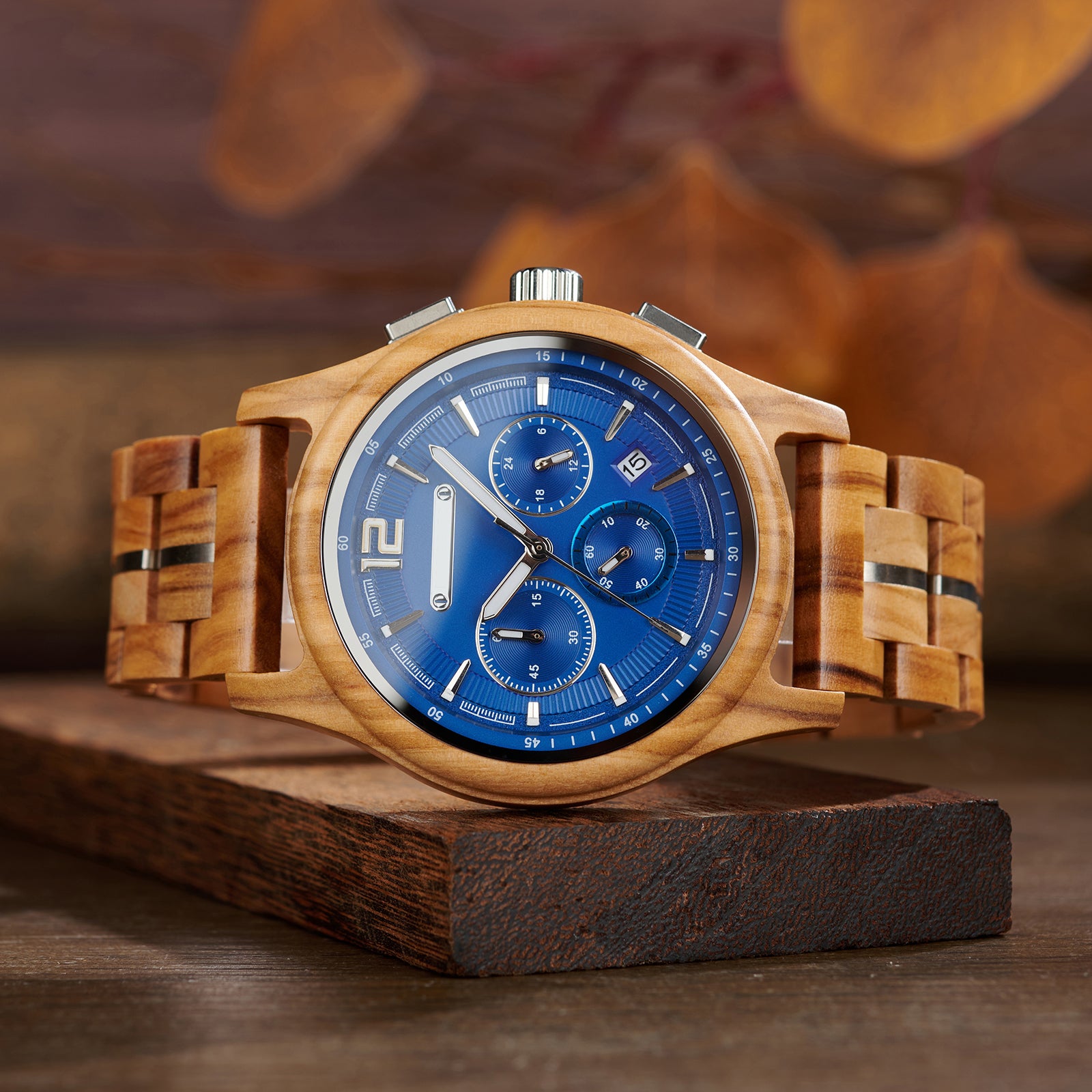A stylish chronograph watch with a blue dial, olive wood case and band (with metal accents), displayed on a wooden block against a warm, blurred background of autumnal leaves.