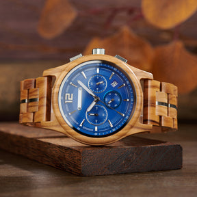 A stylish chronograph watch with a blue dial, olive wood case and band (with metal accents), displayed on a wooden block against a warm, blurred background of autumnal leaves.