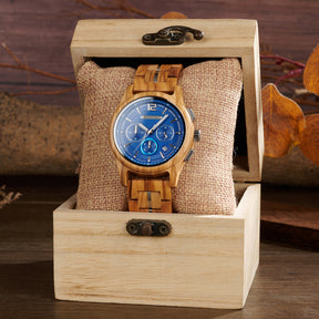 A chronograph watch with a blue dial and olive wood band, nestled on a burlap pillow inside a wooden box, with autumnal leaves and a warm wooden background.