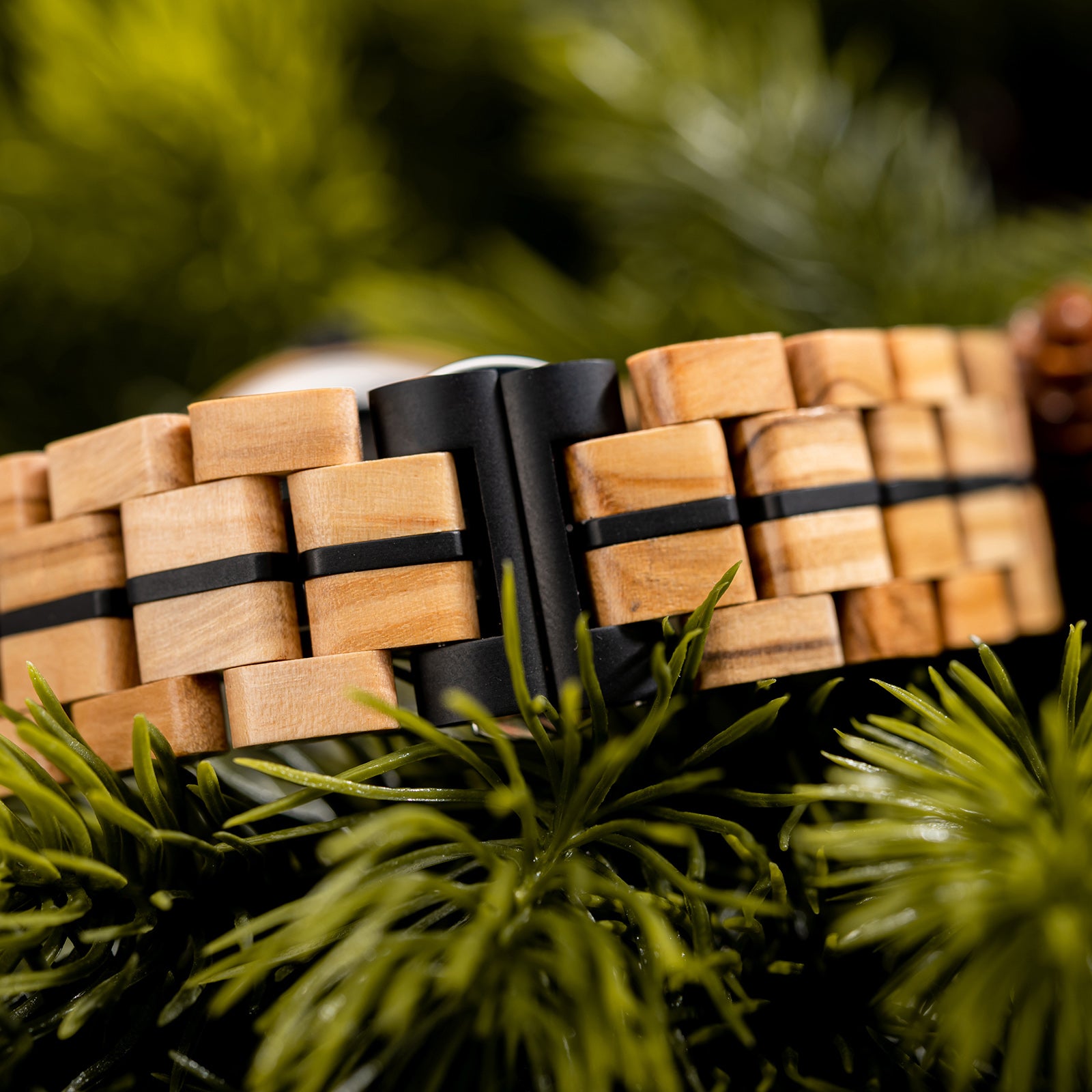 Close-up of a wooden watch band with black metal accents and clasp, placed on green pine foliage.