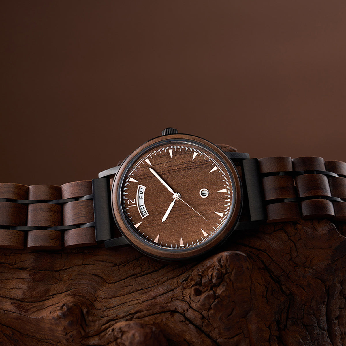 A wooden wristwatch with a brown dial, displaying the day "FRI" and time, features a band made of wooden links and black metal accents, resting on a textured piece of tree bark against a brown background.