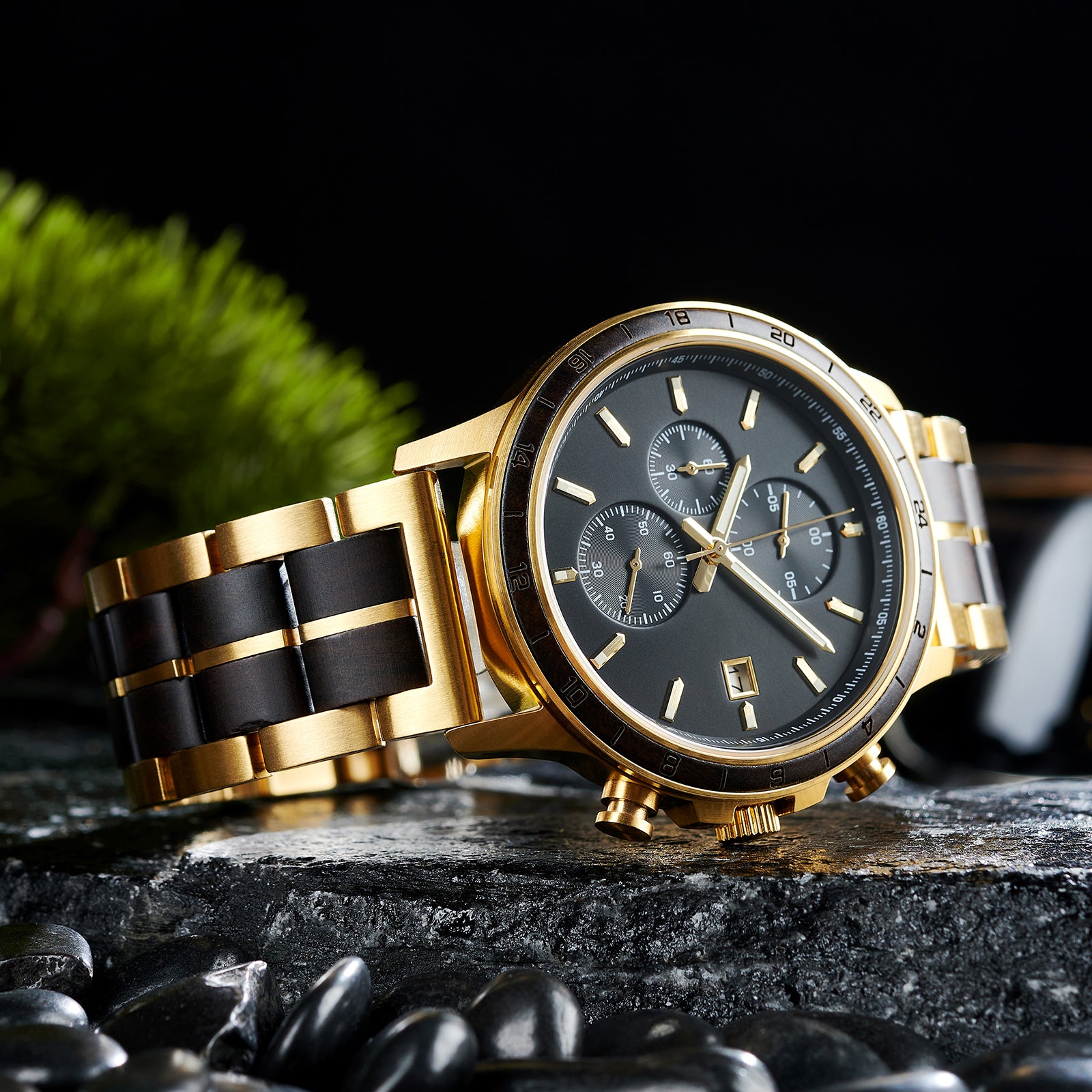 An angled close-up of a chronograph watch with a combination of gold metal and dark wood bracelet, featuring a black dial with multiple sub-dials, 24-hour markers on the bezel, and a date window, placed on wet rock with black pebbles and green foliage in the background.