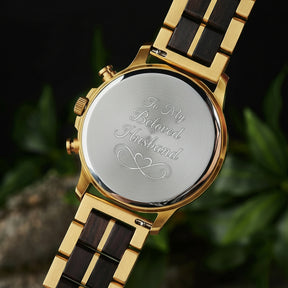 A close-up of the case back of a chronograph watch with a combination of gold metal and dark wood bracelet, showcasing its polished finish, set against green foliage in the background.