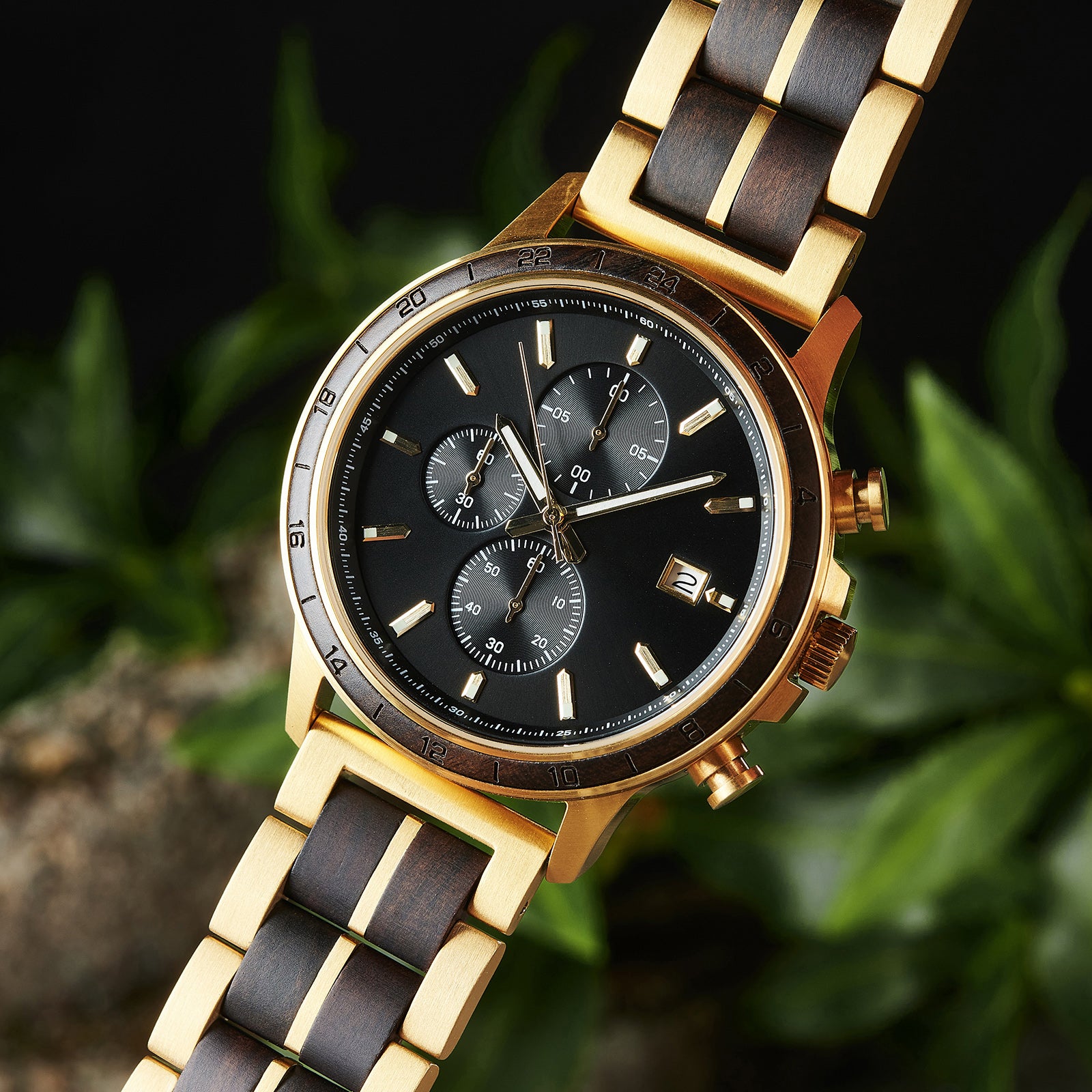 A close-up of a chronograph watch with a combination of gold metal and dark wood bracelet, featuring a black dial with multiple sub-dials, 24-hour markers on the bezel, and a date window, set against green foliage in the background.