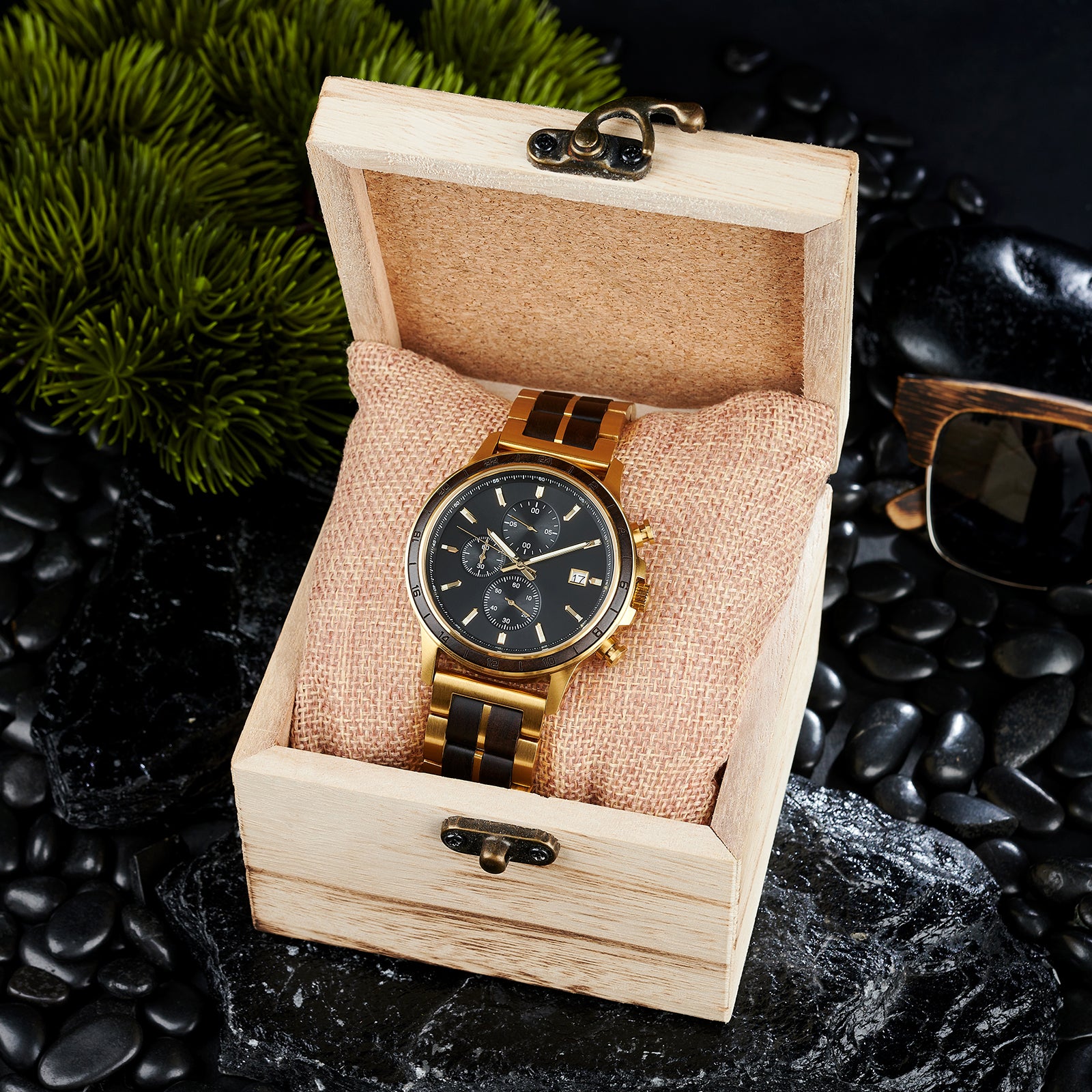 A chronograph watch with a combination of gold metal and dark wood bracelet, featuring a black dial with multiple sub-dials, 24-hour markers on the bezel, and a date window, placed inside a wooden box with a burlap pillow, surrounded by black rocks and green foliage.