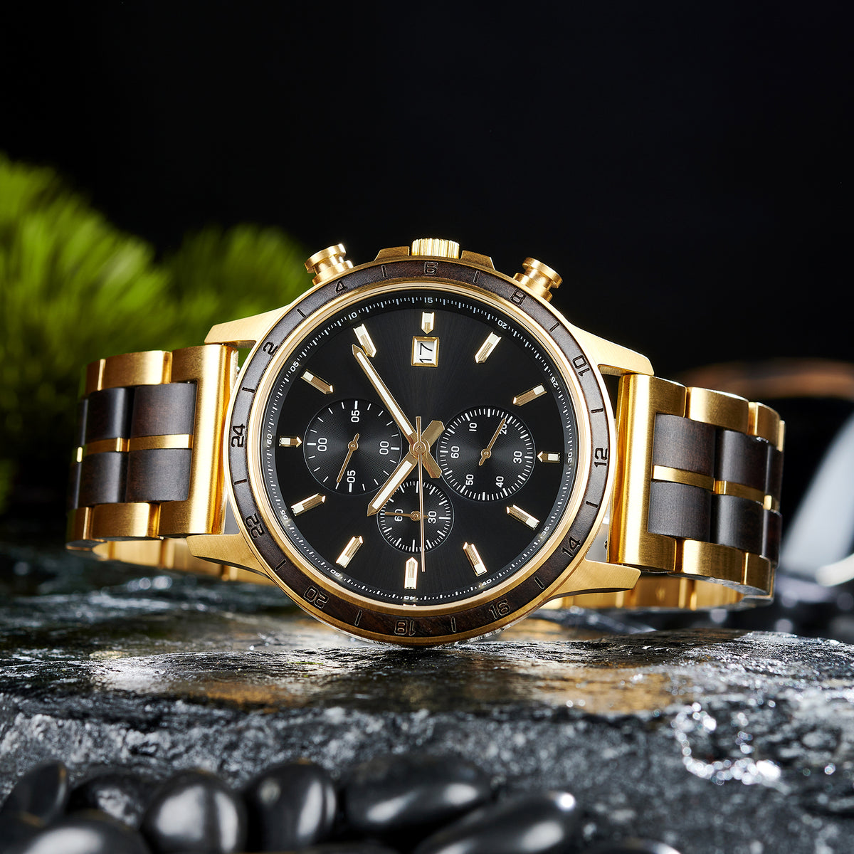 A chronograph watch with a combination of gold metal and dark wood bracelet, featuring a black dial with multiple sub-dials, 24-hour markers on the bezel, and a date window, placed on wet rock with black pebbles and green foliage in the background.