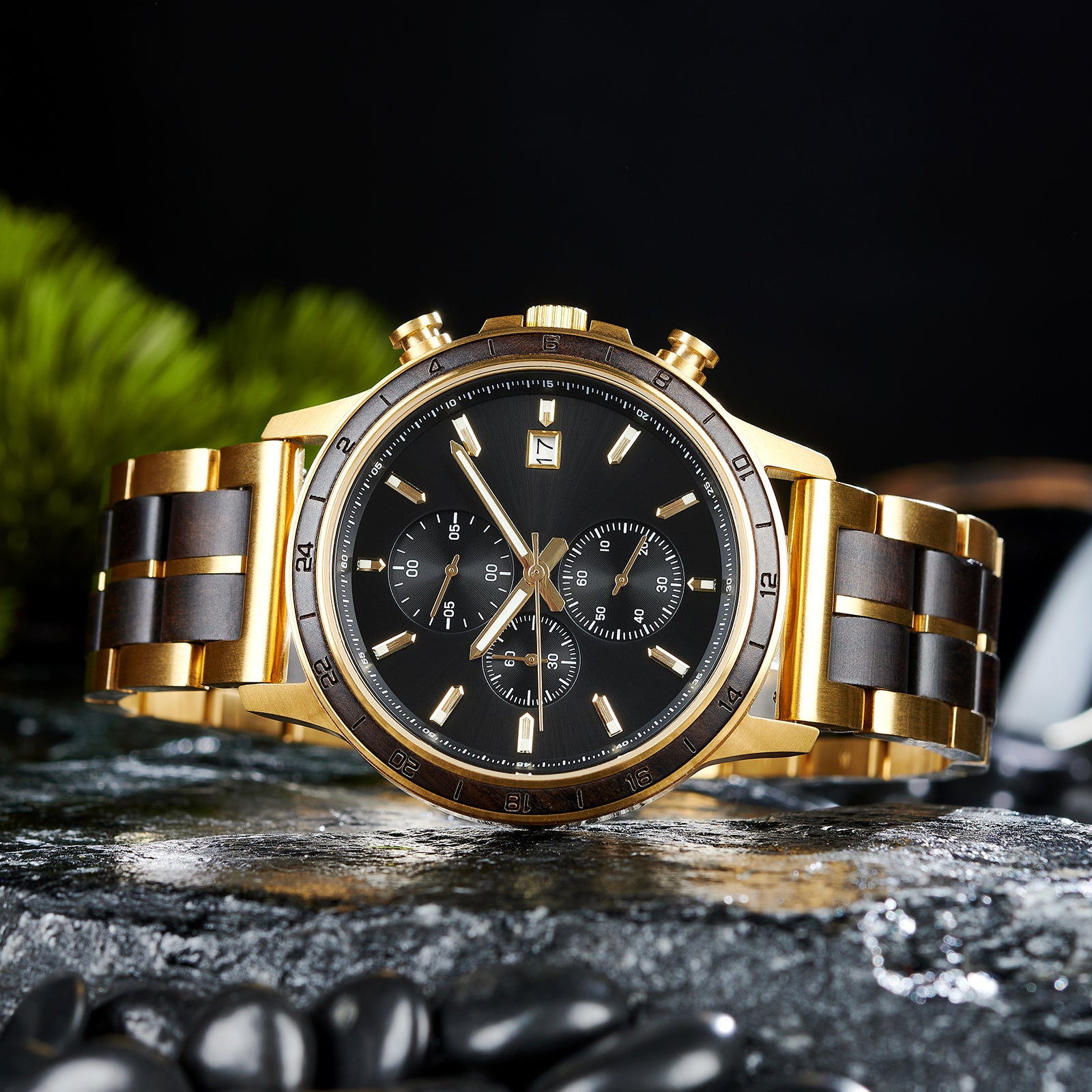 A chronograph watch with a combination of gold metal and dark wood bracelet, featuring a black dial with multiple sub-dials, 24-hour markers on the bezel, and a date window, placed on wet rock with black pebbles and green foliage in the background.
