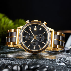 A chronograph watch with a combination of gold metal and dark wood bracelet, featuring a black dial with multiple sub-dials, 24-hour markers on the bezel, and a date window, placed on wet rock with black pebbles and green foliage in the background.