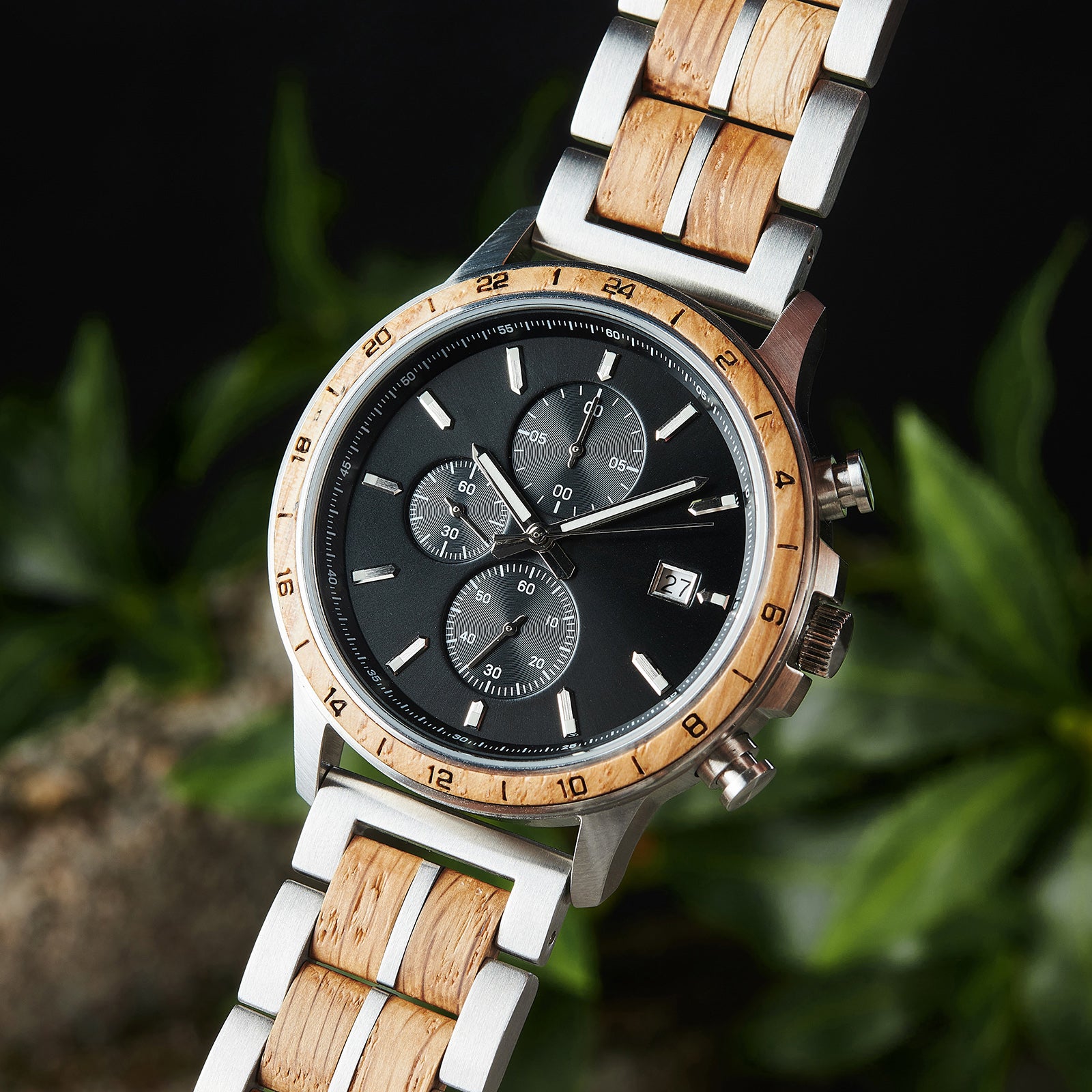 An angled close-up of a chronograph watch with a combination of silver metal and light wood bracelet, featuring a black dial with multiple sub-dials, 24-hour markers on the bezel, and a date window, set against green foliage in the background.