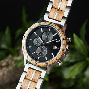 An angled close-up of a chronograph watch with a combination of silver metal and light wood bracelet, featuring a black dial with multiple sub-dials, 24-hour markers on the bezel, and a date window, set against green foliage in the background.