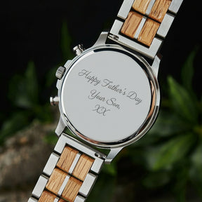 A close-up of the case back of a chronograph watch with a combination of silver metal and light wood bracelet, showcasing its polished finish, set against green foliage in the background.
