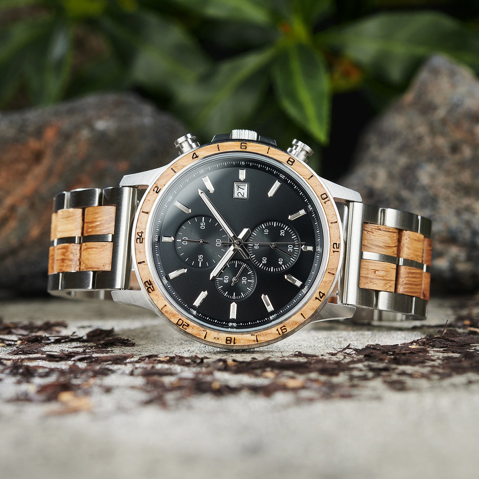 A close-up of a chronograph watch with a combination of silver metal and light wood bracelet, featuring a black dial with multiple sub-dials, 24-hour markers on the bezel, and a date window, placed on rock with green foliage in the background.
