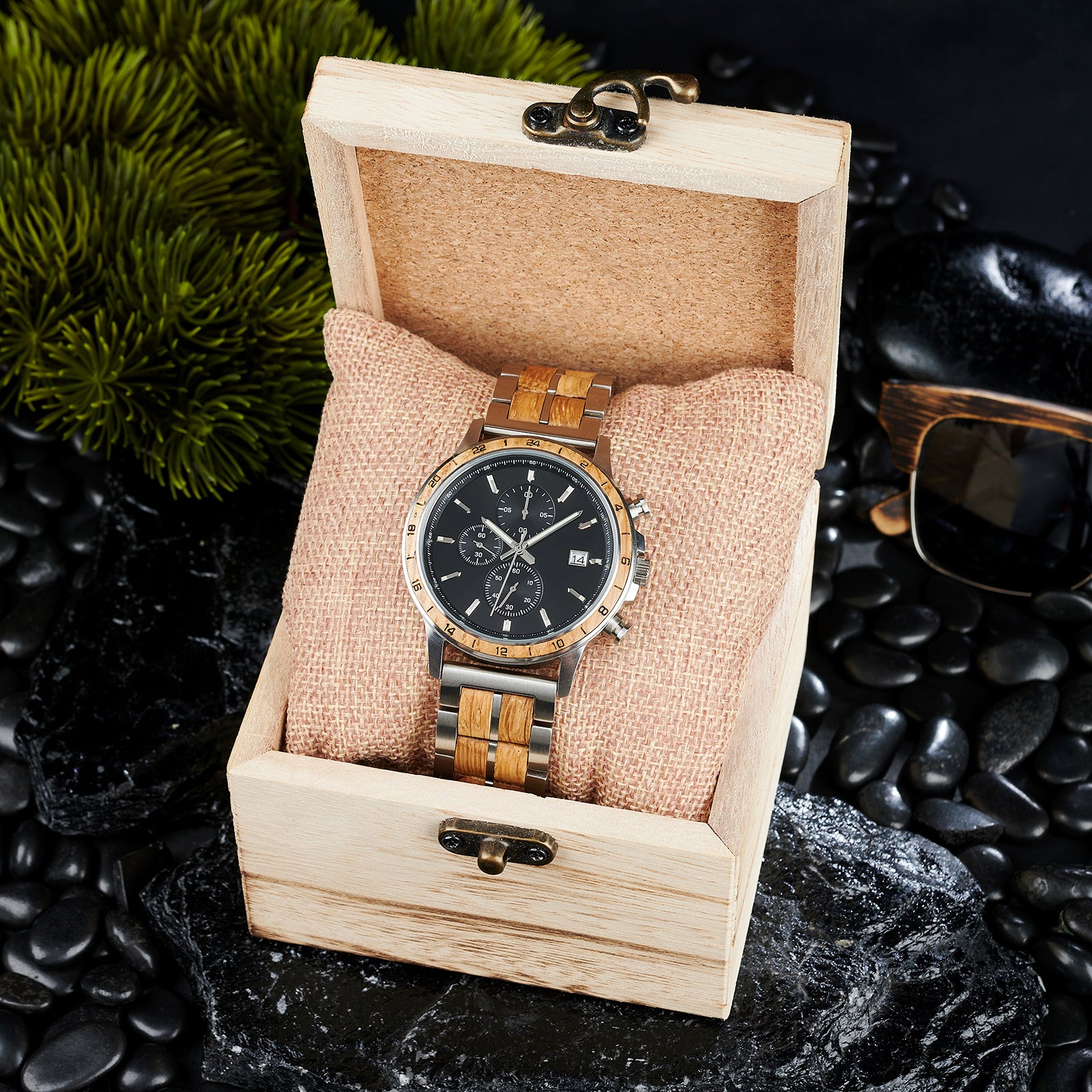 A chronograph watch with a combination of silver metal and light wood bracelet, featuring a black dial with multiple sub-dials, 24-hour markers on the bezel, and a date window, placed inside a wooden box with a burlap pillow, surrounded by black rocks and green foliage.