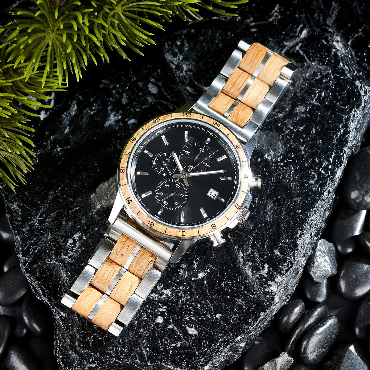 A chronograph watch with a combination of silver metal and light wood bracelet, featuring a black dial with multiple sub-dials, numerical markers on the bezel, and a date window, placed on black rock with green foliage in the background.