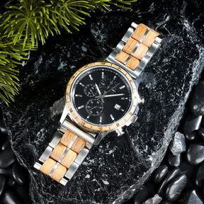 A chronograph watch with a combination of silver metal and light wood bracelet, featuring a black dial with multiple sub-dials, numerical markers on the bezel, and a date window, placed on black rock with green foliage in the background.