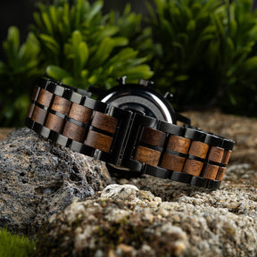 The case back of a chronograph watch with a combination of wooden and black metal bracelet, featuring an engraved Greek key pattern, placed on volcanic rock with green foliage in the background.