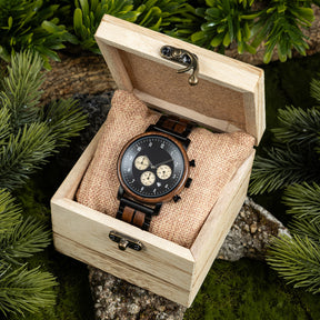 A chronograph watch with a combination of wooden and black metal bracelet, featuring a black dial with beige chronograph sub-dials, numerical hour markers, and a date window, placed inside a wooden box with a burlap pillow, surrounded by green foliage and natural rock elements.