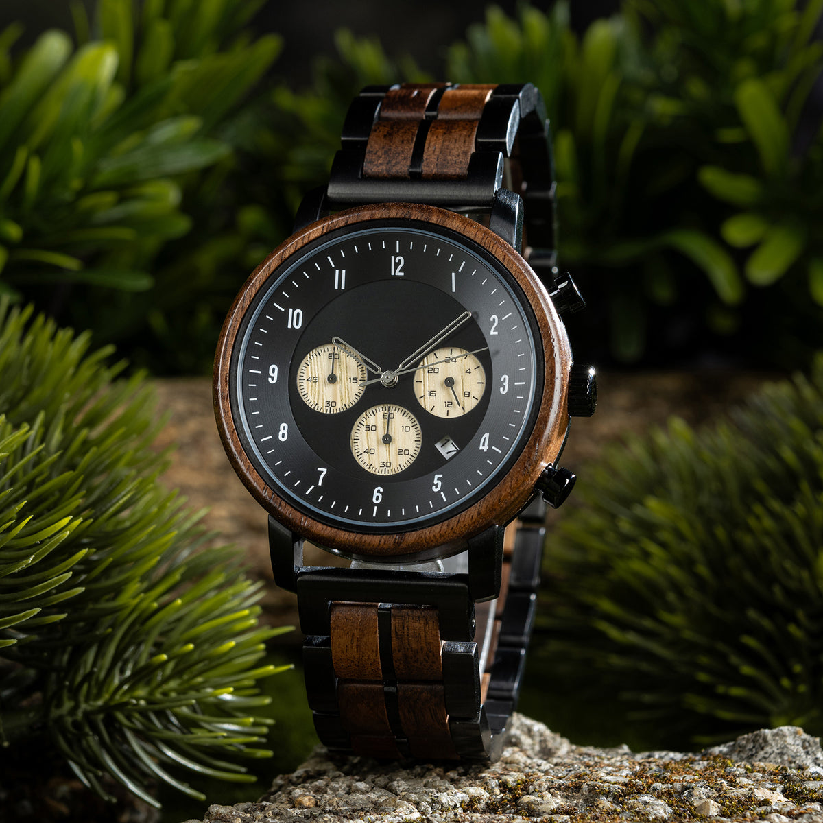 A chronograph watch with a combination of wooden and black metal bracelet, featuring a black dial with beige chronograph sub-dials, numerical hour markers, and a date window, placed on a rock surrounded by green foliage.