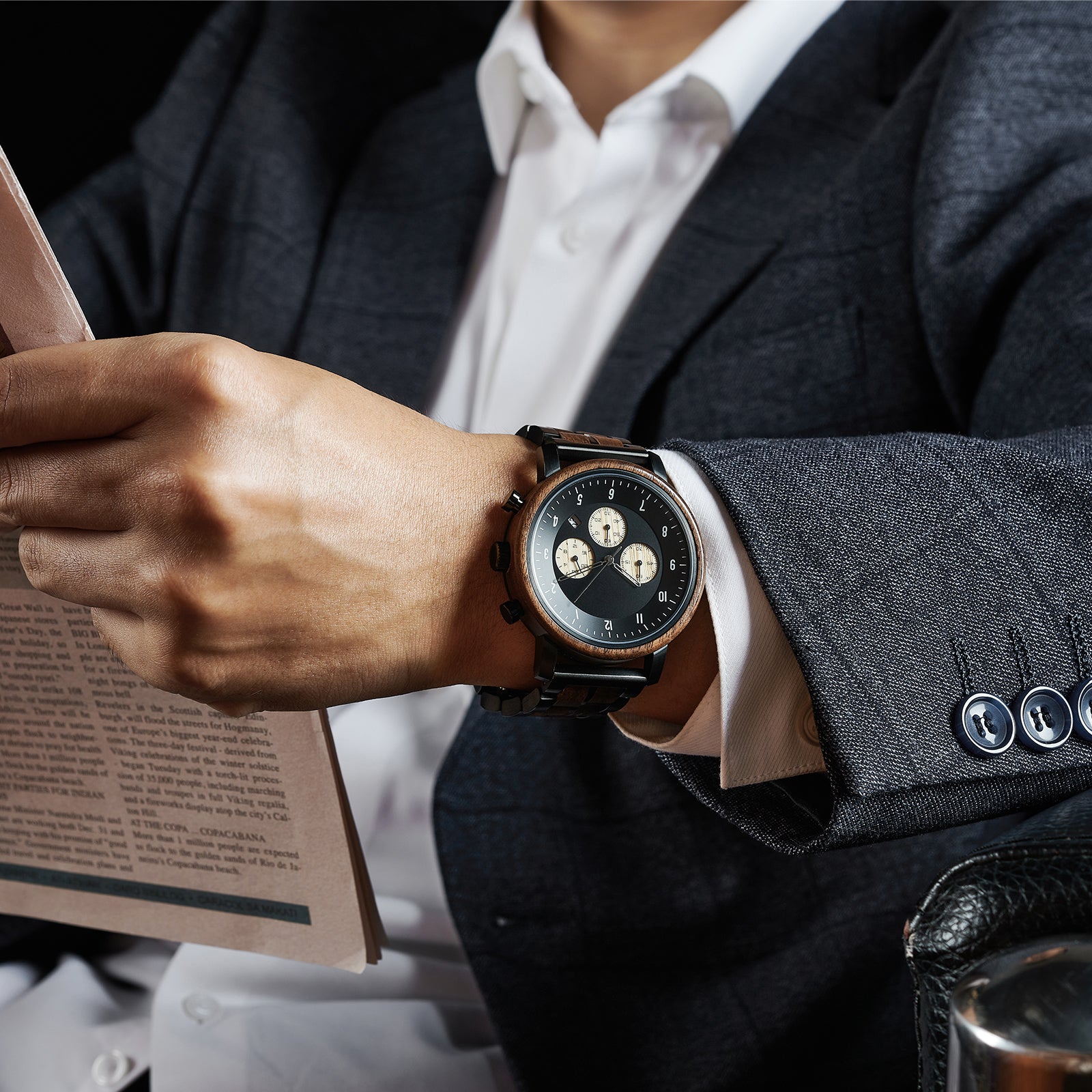 A man wearing a dark suit and white shirt, showcasing a chronograph watch with a combination of wooden and black metal bracelet on his wrist while reading a newspaper, highlighting the watch's sophisticated design.