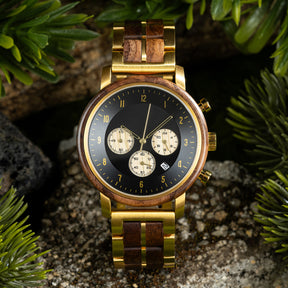 A close-up of a chronograph watch with a combination of wooden and gold metal bracelet, featuring a black dial with gold accents, beige chronograph sub-dials, numerical hour markers, and a date window, placed on a rock with green foliage in the background.