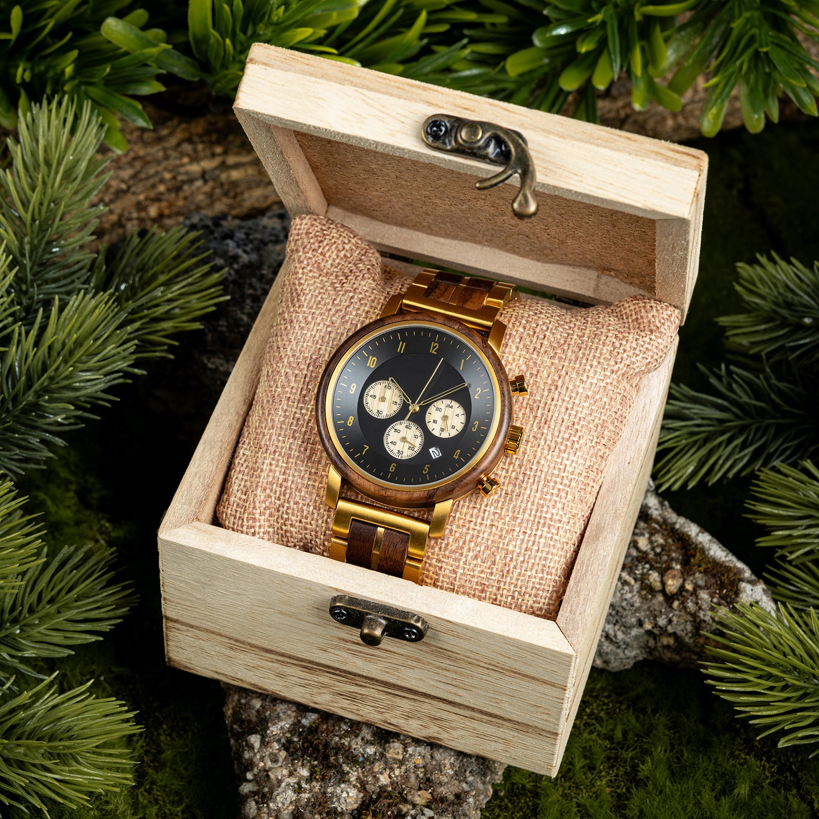 A chronograph watch with a combination of wooden and gold metal bracelet, featuring a black dial with beige chronograph sub-dials, numerical hour markers, and a date window, placed inside a wooden box with a burlap pillow, surrounded by green foliage and natural rock elements.