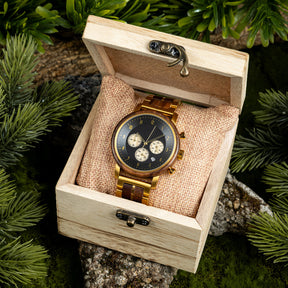 A chronograph watch with a combination of wooden and gold metal bracelet, featuring a black dial with beige chronograph sub-dials, numerical hour markers, and a date window, placed inside a wooden box with a burlap pillow, surrounded by green foliage and natural rock elements.