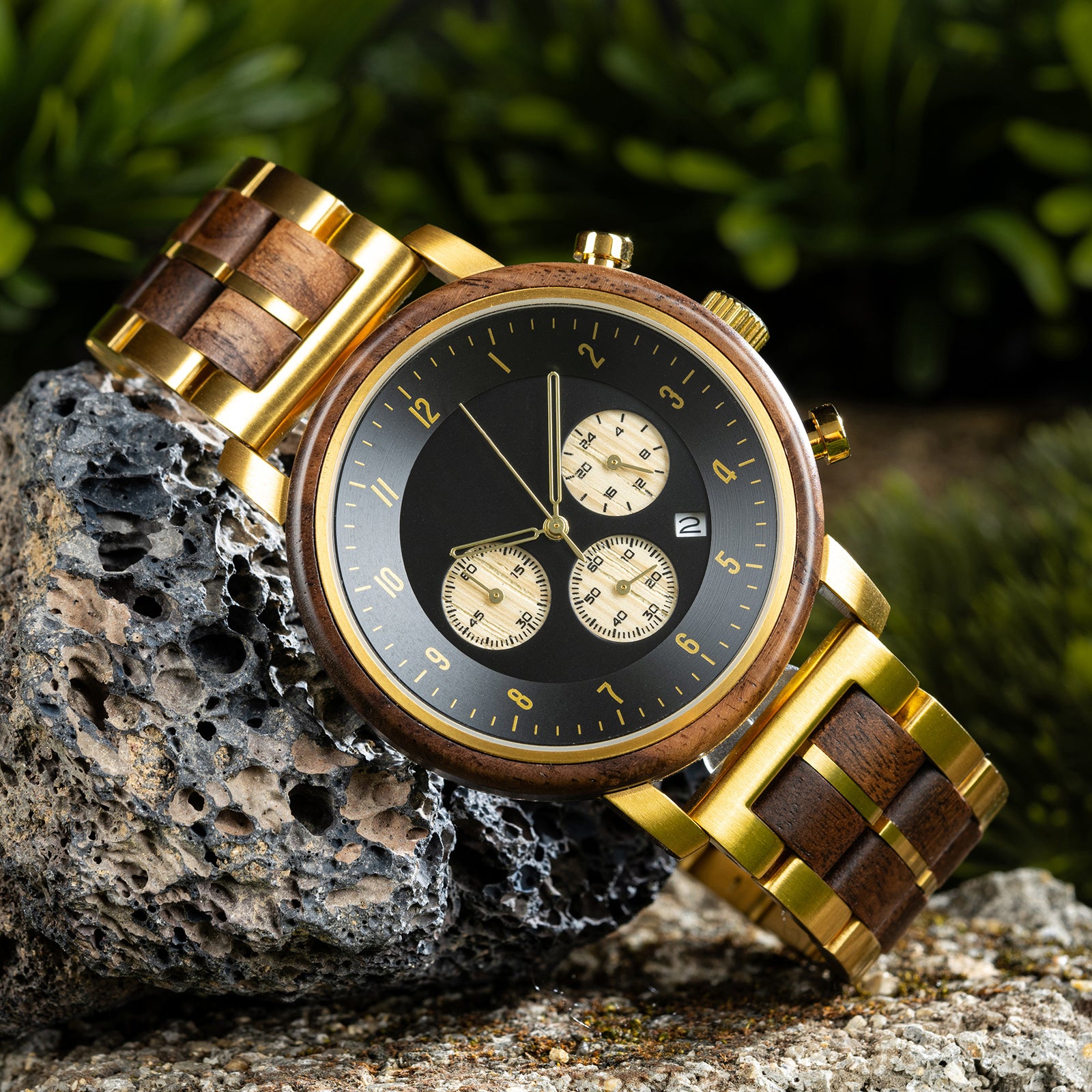 A chronograph watch with a combination of wooden and gold metal bracelet, featuring a black dial with gold accents, beige chronograph sub-dials, numerical hour markers, and a date window, placed on a piece of volcanic rock surrounded by green foliage.