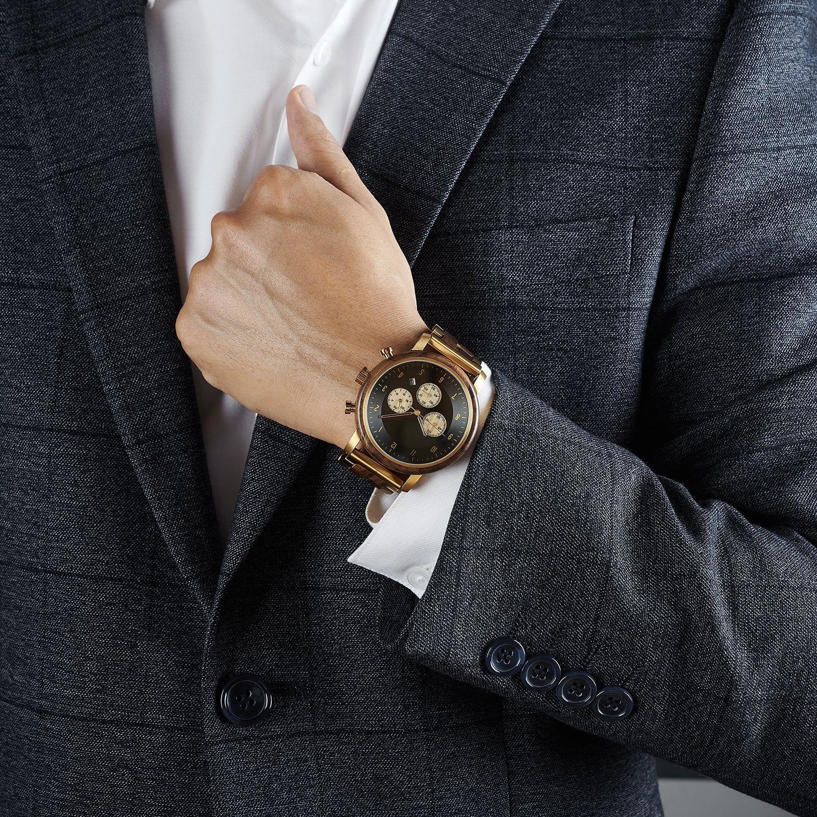 A man wearing a grey suit and white shirt, showcasing a chronograph watch with a combination of wooden and gold metal bracelet on his wrist, featuring a black dial with beige chronograph sub-dials and numerical hour markers, against a neutral background.