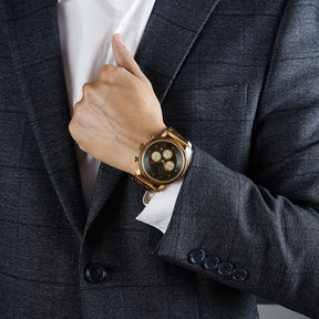 A man wearing a grey suit and white shirt, showcasing a chronograph watch with a combination of wooden and gold metal bracelet on his wrist, featuring a black dial with beige chronograph sub-dials and numerical hour markers, against a neutral background.
