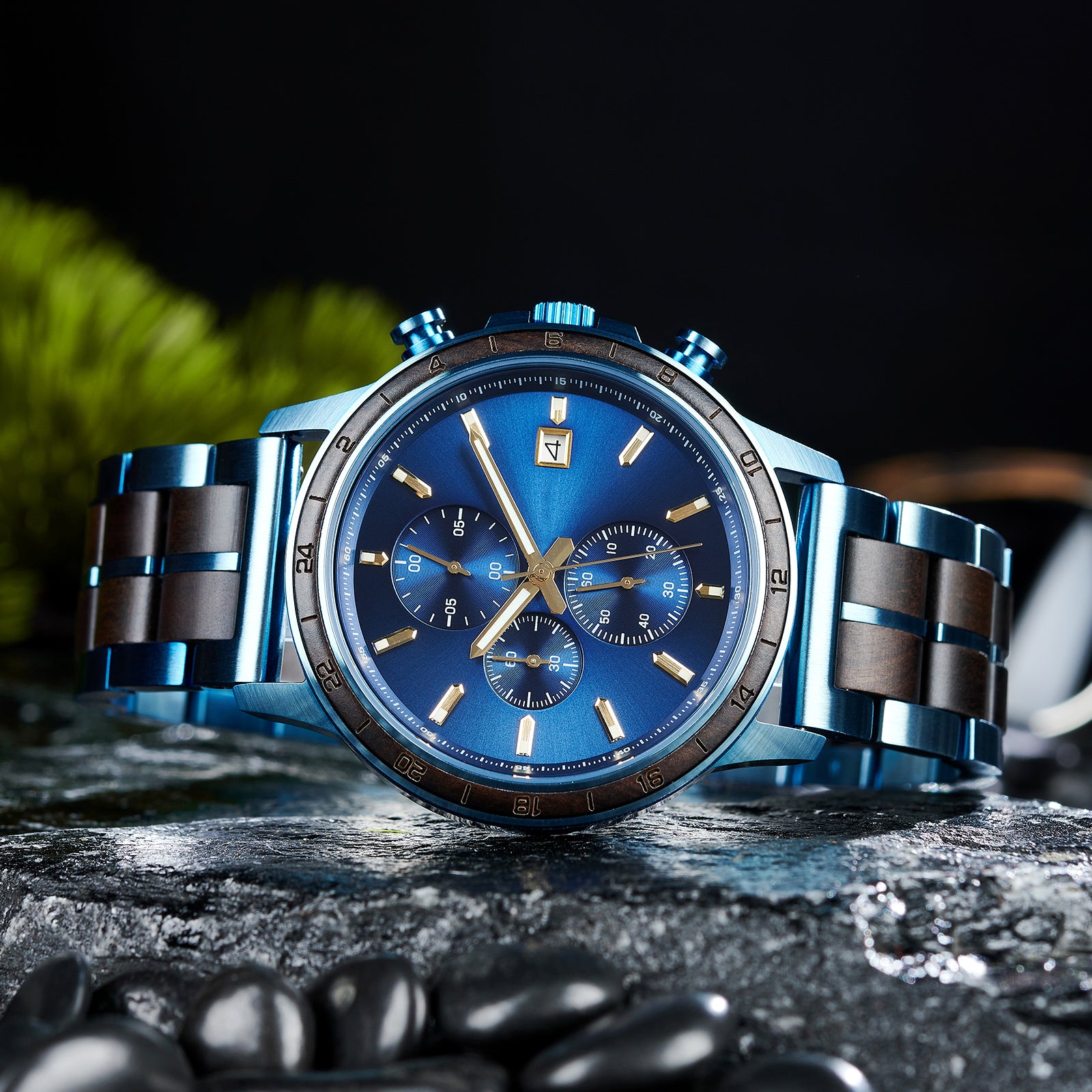 An angled close-up of a chronograph watch with a combination of blue metal and dark wood bracelet, featuring a blue dial with multiple sub-dials, 24-hour markers on the bezel, and a date window, placed on wet rock with black pebbles and green foliage in the background.