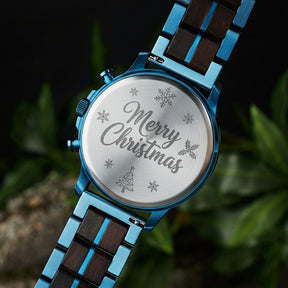 A close-up of the case back of a chronograph watch with a combination of blue metal and dark wood bracelet, showcasing its polished finish, set against green foliage in the background.