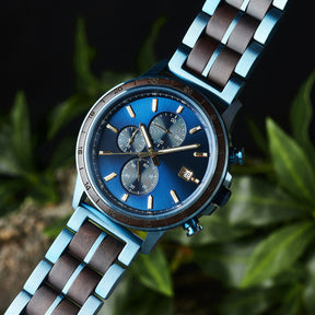 A close-up of a chronograph watch with a combination of blue metal and dark wood bracelet, featuring a blue dial with multiple sub-dials, 24-hour markers on the bezel, and a date window, set against green foliage in the background.