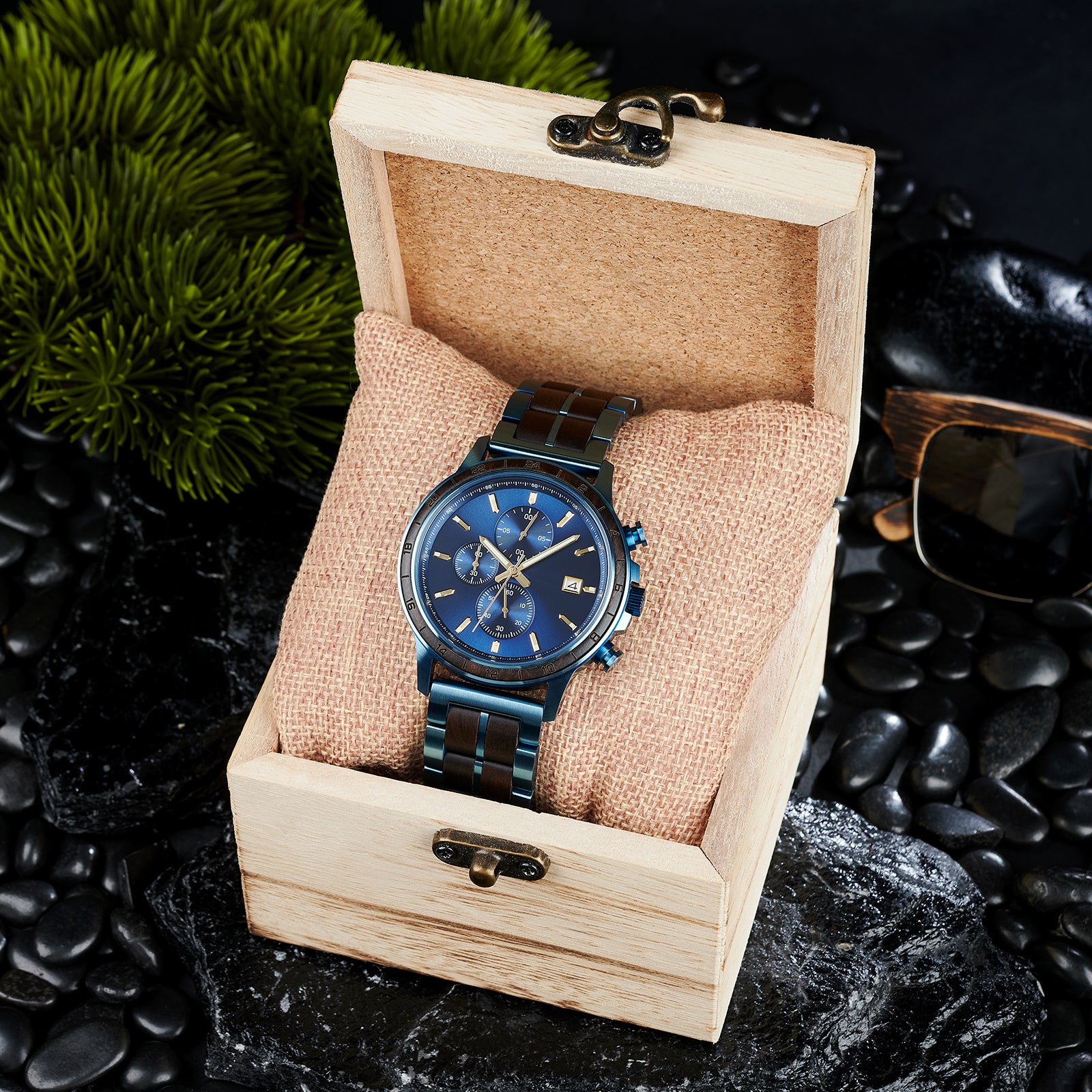 A chronograph watch with a combination of blue metal and dark wood bracelet, featuring a blue dial with multiple sub-dials, 24-hour markers on the bezel, and a date window, placed inside a wooden box with a burlap pillow, surrounded by black rocks and green foliage.