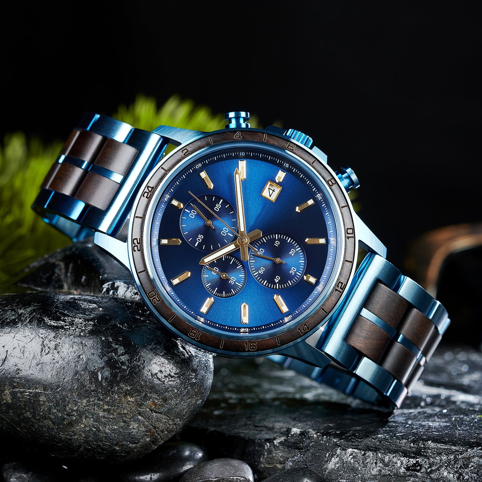 A chronograph watch with a combination of blue metal and dark wood bracelet, featuring a blue dial with multiple sub-dials, 24-hour markers on the bezel, and a date window, placed on wet rock with black pebbles and green foliage in the background.