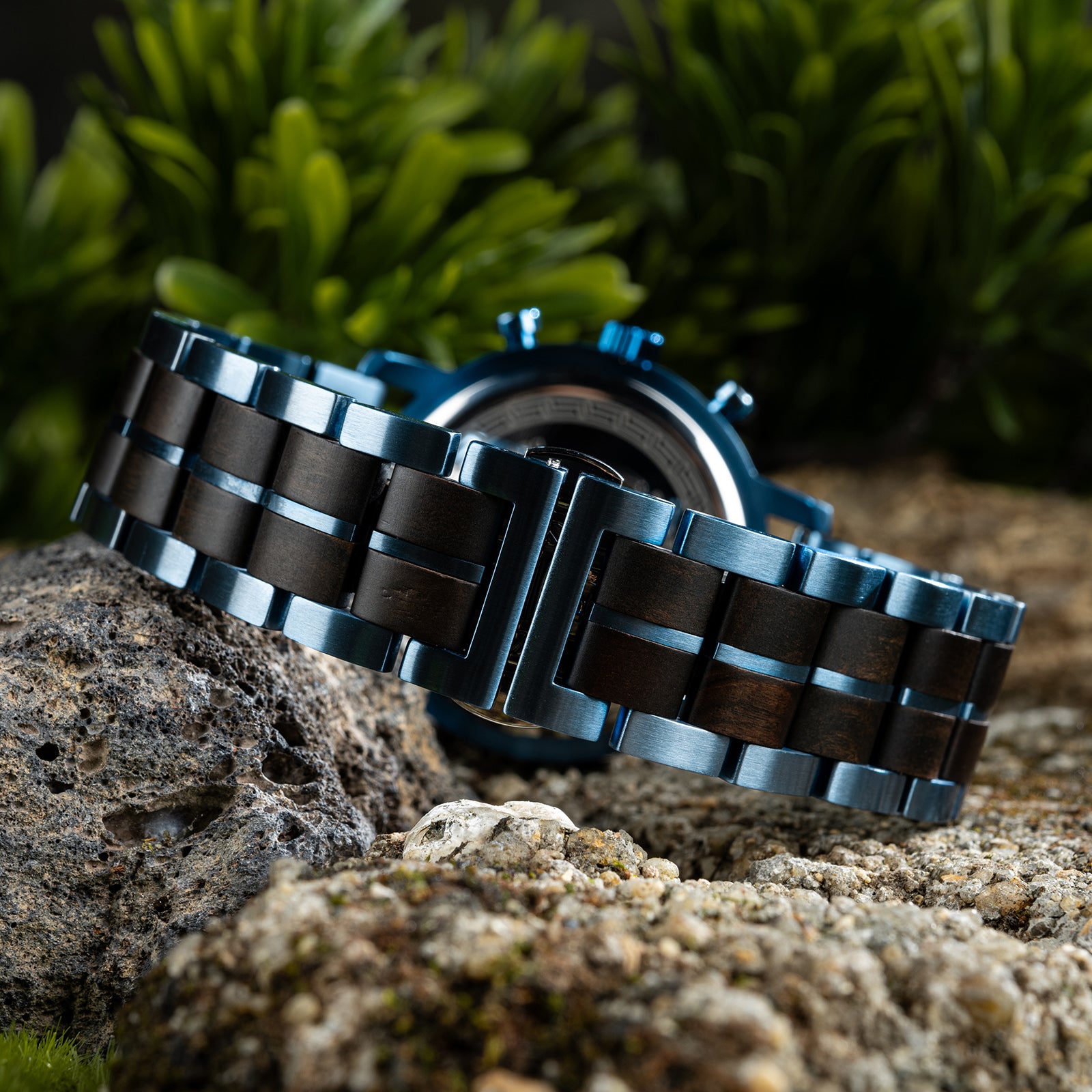 The case back of a chronograph watch with a combination of wooden and blue metal bracelet, featuring an engraved Greek key pattern, placed on volcanic rock with green foliage in the background.