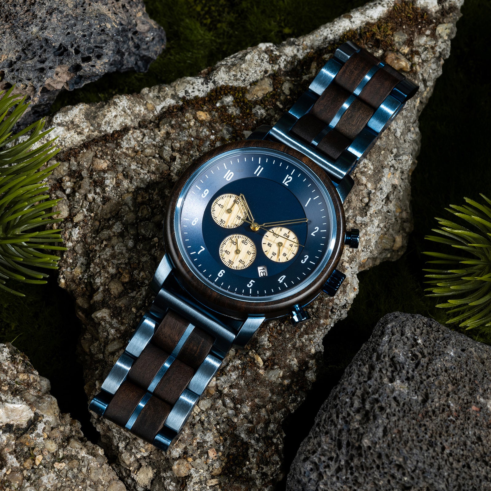 A close-up of a chronograph watch with a combination of wooden and blue metal bracelet, featuring a blue dial with beige chronograph sub-dials, numerical hour markers, and a date window, placed on volcanic rock with green foliage in the background.
