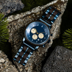 A close-up of a chronograph watch with a combination of wooden and blue metal bracelet, featuring a blue dial with beige chronograph sub-dials, numerical hour markers, and a date window, placed on volcanic rock with green foliage in the background.
