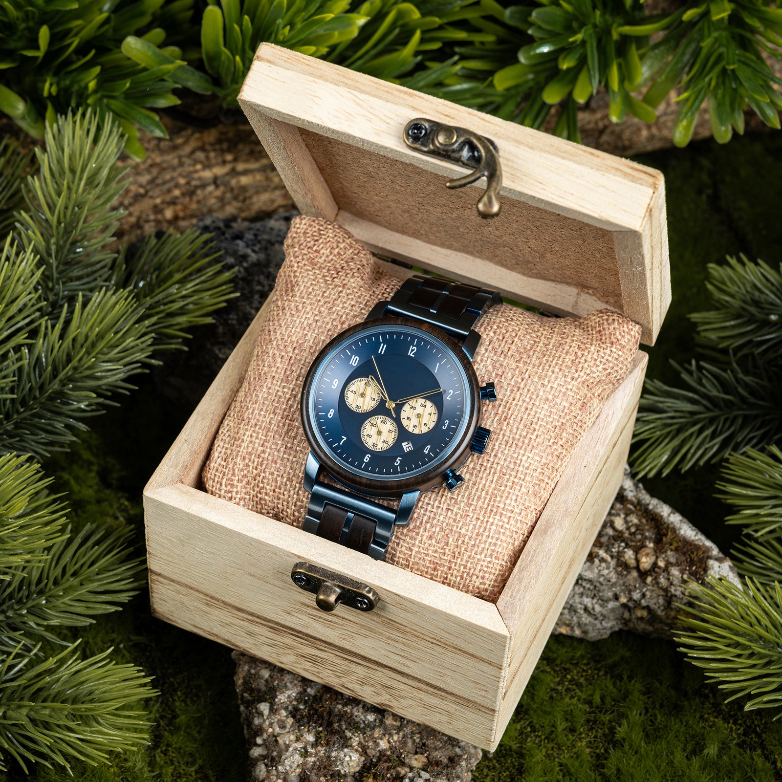 A chronograph watch with a combination of wooden and blue metal bracelet, featuring a blue dial with beige chronograph sub-dials, numerical hour markers, and a date window, placed inside a wooden box with a burlap pillow, surrounded by green foliage and natural rock elements.