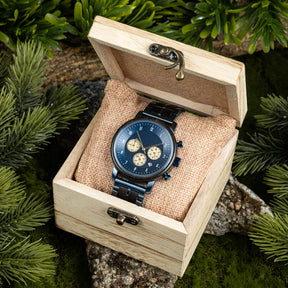 A chronograph watch with a combination of wooden and blue metal bracelet, featuring a blue dial with beige chronograph sub-dials, numerical hour markers, and a date window, placed inside a wooden box with a burlap pillow, surrounded by green foliage and natural rock elements.